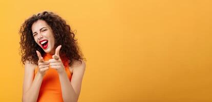 Hey what is up. Friendly and outgoing attractive curly-haired female with red lipstick pointing at camera with finger guns as if greeting or hinting friend winking at camera over orange background photo