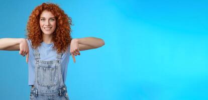 Charismatic sassy flirty redhead daring ginger girl curly haircut pointing down index fingers smiling broadly enthusiastic explore new store pointing promo like cool advertisement blue background photo