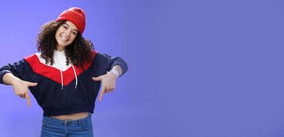 Waist-up shot of cool and stylish young woman with curly hair smiling flirty and joyful pointing down showing promotion as tilting head happily and posing over blue background in outdoor clothes photo