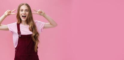 Girl staying positive lifting after falling. Charming carefree friendly-looking young woman with small scar on arm and tattoo showing peace gestures, open mouth and looking at camera over pink wall photo