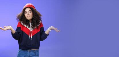 Not getting clue. Cute silly woman with curly hair shrugging with pursed lips and unbothered expression holding hands sideways looking right being unaware and confused, uncertain what answer photo