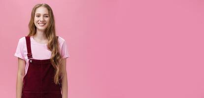 Portrait of charming slim young girl with long fair hair in corduroy overalls smiling joyfully standing upright with positive grin posing against pink background happy and friendly photo