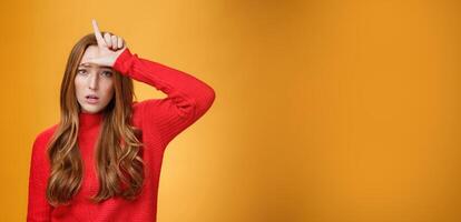 Arrogant and self-satisfied redhead woman humiliating person showing loser sign on forehead mocking and disdain rival standing confident and snobbish over orange background photo