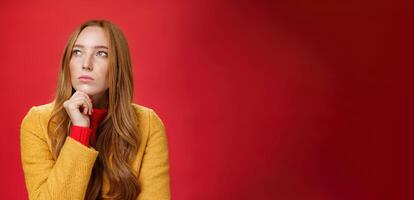 Close-up shot of determined and focused creative thoughtful redhead female looking at upper left corner touching chin, thinking, making choice or remembering information over red background photo