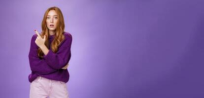 Astonished excited redhead woman open mouth and raise eyebrows questioned as seing shocking thing pointing at upper left corner, asking question impressed and surprised over purple wall photo