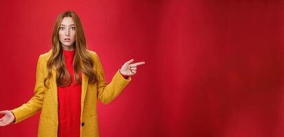 Confused and questioned cute redhead girl in yellow autumn coat pointing right as shrugging making clueless gesture with hand open mouth wondered as asking question over red background photo