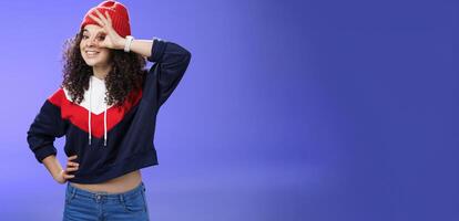 Okay girl agrees. Friendly-looking happy and charming young curly-haired girl in warm beanie showing ok gesture or circle over eye as peeking at camera and smiling broadly with joy over blue wall photo