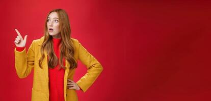 Portrait of empathic shocked and surprised stunned redhead woman in yellow coat open mouth amazed and speechless pointing and looking at upper left corner concerned and shook over red wall photo