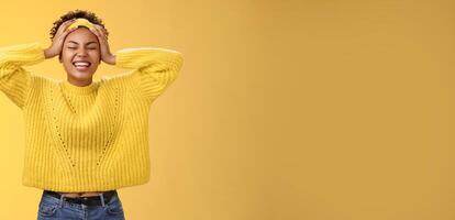 Carefree happy lucky african girl in sweater headband touching head having fun smiling relieved excited laughing out loud close eyes rejoicing spend good time partying, standing yellow background photo