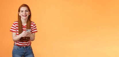 Carefree attractive woman red long hair laughing amused, having fun, chatting friend, hold smartphone, look camera sincere rejoice smile, texting joke, found funny meme internet, orange background photo