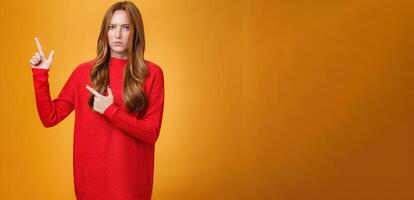 Intense suspicious and doubtful european ginger girl in warm sweater frowning hesitant and confused pointing at upper left corner asking question, looking with disbelief over orange background photo