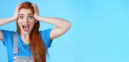 Shocked redhead girl shouting panic, feel pressured deadlines, scared see mess, grab head, scream frightened distressed, stare camera hopeless, anxiously react losing lottery, blue background photo