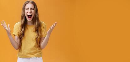 Studio shot of young woman during argument losing temper standing pressured and pissed yelling out loud from hate and anger gesturing with raised palms making furious face posing against orange wall photo