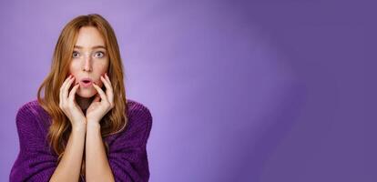 Shocked redhead female friend touching face and open mouth as looking with widen eyes at camera, being overwhelmed with shook and amazement hearing terrible story over purple background photo