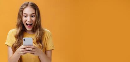Excited and thrilled charismatic young happy girl in yellow t-shirt smiling with opened from joy mouth holding smartphone staring at cellphone screen astonished carried away with cool phone game photo