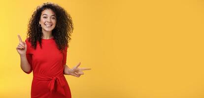 Charismatic cute and happy young curly-haired european female in red dress dancing and pointing up and right, showing sideways as presenting choices picking, making decision and smiling at camera photo