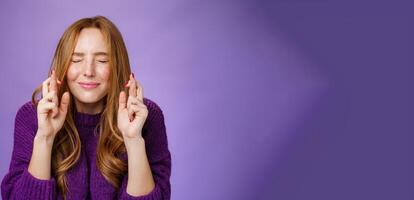Hopeful and dreamy cute tender redhead 20s girl in purple sweater making wish faithfully close eyes and cross fingers for good luck as anticipating miracle happen, supplicating over violet wall photo