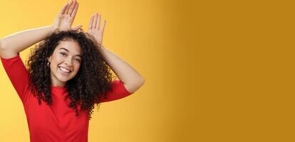 Close-up shot of charismatic playful and tender young kind woman with curly hair playing having fun showing bunny ears with hands on head tilting head joyfully fooling around and smiling at camera photo