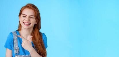 Cheerful carefree redhead woman sincere smile, pointing left introduce copy space joyfully close eyes, show white toothy grin delighted, feel upbeat rejoicing, joyful positive mood, blue background photo