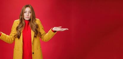 Who new it happen. Innocent and cute redhead unaware girl with freckles in yellow coat shrugging with hands spread sideways, pouting sad and gloomy, being uanware and clueless over red wall photo