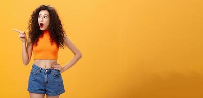 Woman peeking at neighbours window being surprised and amazed pointing and staring speechless left standing over orange background with dropped jaw intrigued and thrilled look in cropped top photo