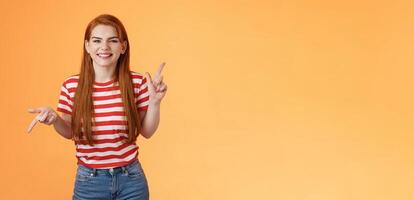 Carefree daring redhead cheerful girl dancing, pointing sideways, showing different sides, left right copy space, laughing and smiling broadly, joyfully recommend cool product, orange background photo