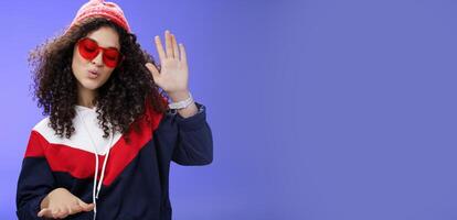 Studio shot of cool and stylish dj girl in red beanie and sunglasses raising palm as enjoying cool music and dancing to rhythm folding lips having fun at awesome party posing chill over blue wall photo