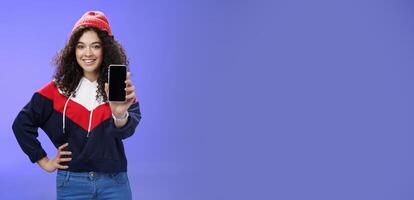Girl showing her post as pulling smartphone screen towards camera smiling delighted waiting opinion, holding hand on waist in confident pose wearing warm winter red beanie and sweatshirt photo