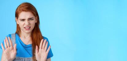 Close-up uninterested displeased ignorant picky redhead girlfriend, frowning make disgusted grimace raise hands block stop gesture, refuse, waving arms no rejection sign, stand blue background photo