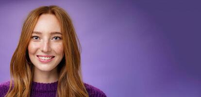 Headshot of charming attractive and happy young redhead woman with freckles and bright white smile, grinning satisfied at camera as posing over purple background friendly and delighted photo