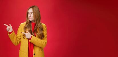 Studio shot of intense suspicious and displeased redhead female in yellow coat pointing and looking serious left squnting and frowning making focused look at something disturbing and doubtful photo