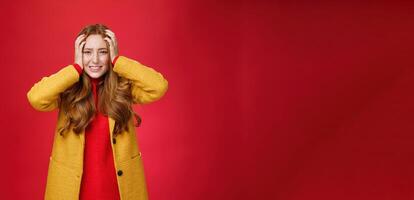 Portrait of redhead woman in panic holding hands pressed to head clenching teeth and frowning puzzled and troubled, standing anxious and concerned freaking out over red background photo