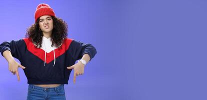 Snobbish and displeased stylish and cool street girl with curly hair in beanie and sweatshirt pointing down grimacing as feeling unsatisfied and disappointed posing against blue background photo