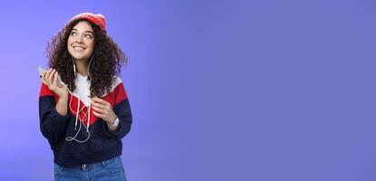 Dreamy charming and happy kind girl recalling nice memories as listening nostalgic song in earphones holding smartphone looking at upper left corner with broad smile over blue background photo