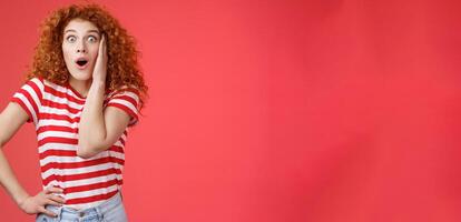Shocked amused emotive redhead ginger girl curly haircut popping eyes stare camera fascinated impressed open mouth gasping entertained react incredible store price offer red background photo