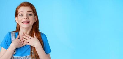 Grateful sincere cute charming redhead female thanking friends, smiling delighted appreciate nice tender gesture, press palms chest heart, grateful, look admiration and love, stand blue background photo