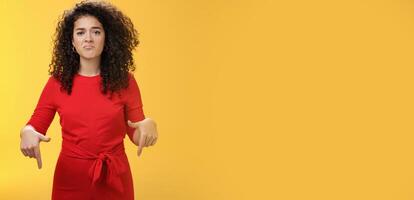 Portrait of gloomy girl with sad face pursing lips down frowning disappointed and pointing down with regret and unsatisfied expression, missing opportunity feeling upset over yellow background photo