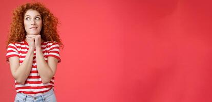 Cheerful intrigued redhead silly curly-haired woman smiling interested hold hands chin face-line imaging thinking about tasty burger look away desire have bite standing red background photo
