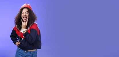 Stylish and cool energized swag girl with curly hairstyle in warm beanie showing rock-n-roll gesture near mouth and sticking out tongue smiling having fun, partying and enjoying life over blue wall photo