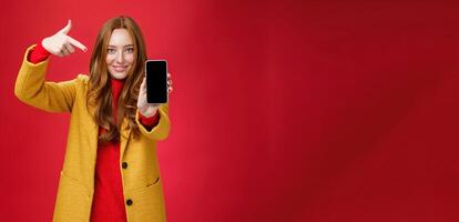 Pick this phone you never regret. Portrait of friendly-looking attractive and confident redhead glamour girlfriend in yellow coat showing smartphone pointing at mobile as smiling broadly at camera photo