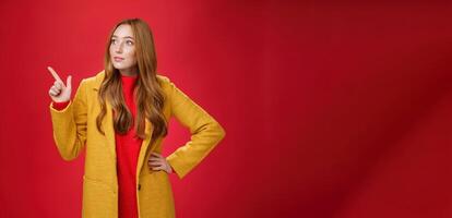 Wondered curious good-looking redhead woman in coat stopping on way looking and pointing at upper left corner curious and interested, observing surprising promotion over red background photo