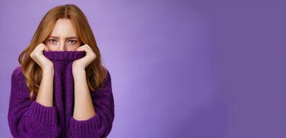 Gloomy upset redhead girlfriend pulling collar of purple sweater on nose, frowning and squinting offended, being aggrieved and resent, sulking moody and displeased over violet background photo