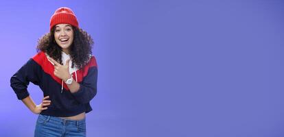 Stylish and sociable friendly-looking attractive woman with curly hair in warm beanie pointing at upper left corner laughing from joy and amazement having fun posing happily over blue background photo