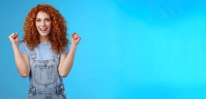 Hooray successful day. Cheerful chrismastic redhead girl summer dungarees clench fists joyfully smiling broadly determined achieve success, triumphing winning game standing upbeat blue background photo