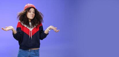 Upset girl cannot get clue what happened shrugging turning away as pouting feeling questioned and unsure what answer, standing clueless and puzzled in sweatshirt and stylish hat over blue background photo
