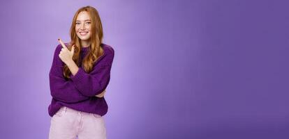 Charming friendly-looking bright redhead woman with freckles and makeup in purple warm sweater pointing at upper left corner and smiling delighted and cute at camera as showing cool place hang out photo