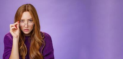 Questioned and unsure cute smart female detective with ginger hair looking suspicious from under forehead taking off glasses and looking at camera doubtful and uncertain over purple background photo