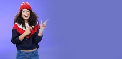 Enthusiastic friendly-looking attractive young 20s woman with curly hair in warm beanie and sweatshirt smiling open mouth amazed and delighted as pointing at upper right corner over blue background photo