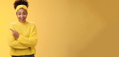 Amused charismatic smiling black cute girl in headband sweater widen eyes drop jaw astonished hear about awesome interesting new place talking friend standing yellow background pointing left amazed photo