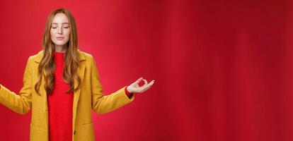 Girl keep calm releasing stress with meditation, posing in yellow coat, close eyes and looking relieved as extending hands sideways with mudra gesture, doing yoga against red background in lotus pose photo